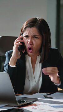 Frustrated office woman talking on the phone while working on her laptop, vertical shot