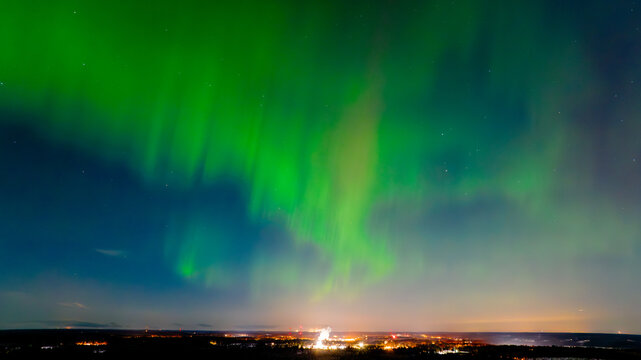 Aerial view of vibrant green aurora borealis dances across the dark sky over the distant twinkling lights of the city, Karis, Uusimaa, Finland.