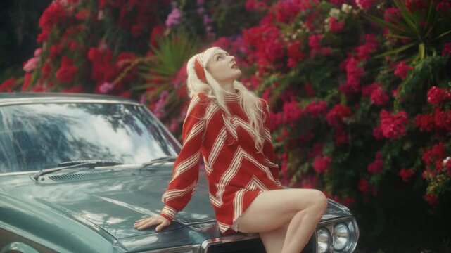Blonde Woman In Retro Attire By Automobile. Serene Woman Resting Near Vehicle Amidst Lush Flowers