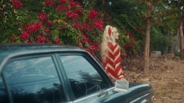 Blonde Woman In Retro Attire By Automobile. Serene Woman Resting Near Vehicle Amidst Lush Flowers