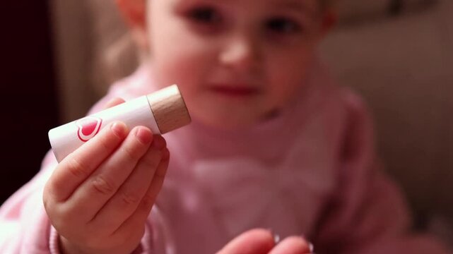 A close-up of a mother's hand holding a wooden toy lipstick and passing it to her little girl in a pink dress. Interaction and shared play at home.