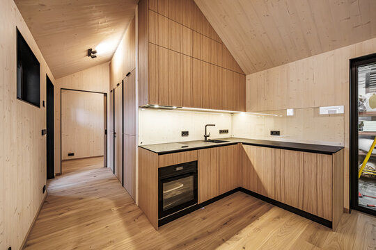 Warm wooden interior with L-shaped kitchen, dark countertops, built-in oven, and sloped ceiling. Ample natural wood flooring provides copy space. No visible branding or logos