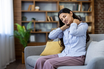 Young indian woman sitting on sofa massaging neck and shoulders, showing cervical ache and muscle...