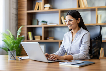 Fototapeta premium Young businesswoman with dark hair and glasses working on her laptop in a modern wooden office, smiling while typing, showing dedication and efficiency in her professional environment