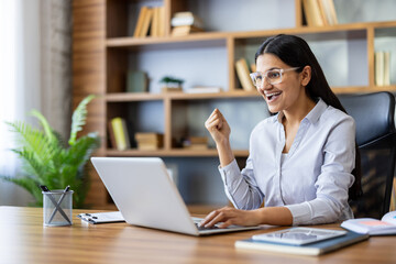 Fototapeta premium Indian woman at laptop pumps fist in celebration of career success and remote work achievement, smiling in a modern home office as a freelancer or entrepreneur completes a win