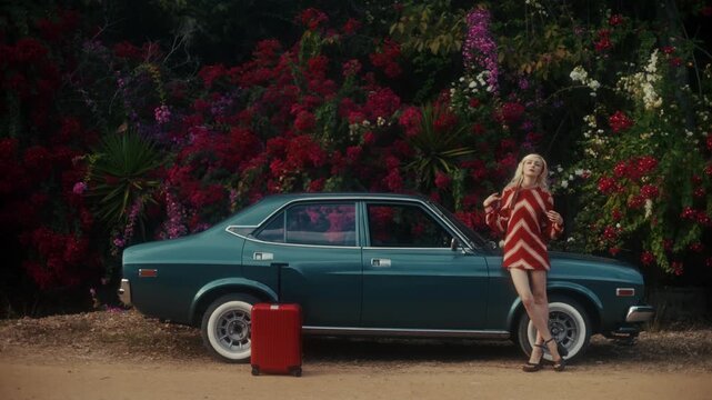 Blonde Woman In Retro Attire By Automobile. Serene Woman Resting Near Vehicle Amidst Lush Flowers
