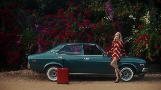 Blonde Woman In Retro Attire By Automobile. Serene Woman Resting Near Vehicle Amidst Lush Flowers
