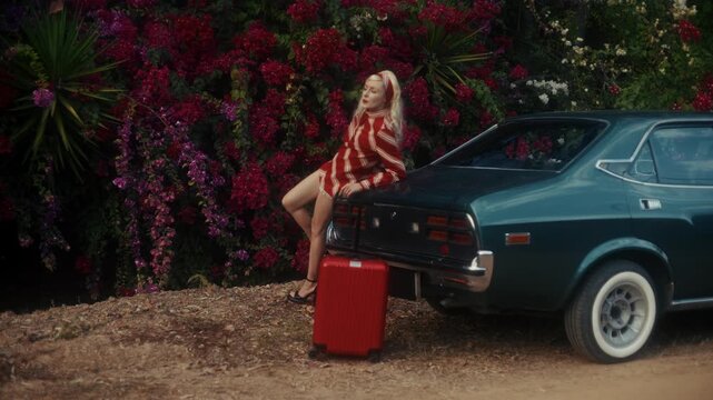 Blonde Woman In Retro Attire By Automobile. Serene Woman Resting Near Vehicle Amidst Lush Flowers