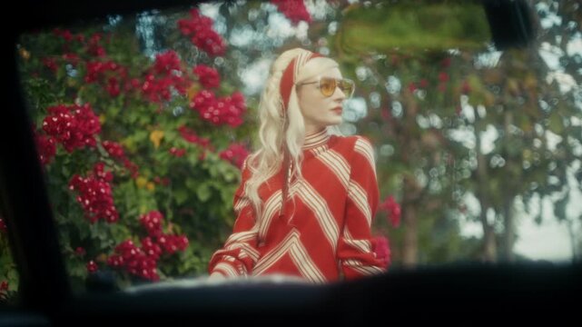 Blonde Woman In Retro Attire By Automobile. Serene Woman Resting Near Vehicle Amidst Lush Flowers