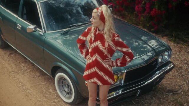 Blonde Woman In Retro Attire By Automobile. Serene Woman Resting Near Vehicle Amidst Lush Flowers