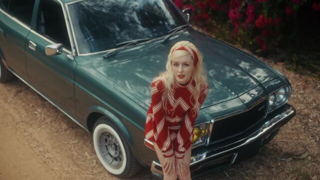 Blonde Woman In Retro Attire By Automobile. Serene Woman Resting Near Vehicle Amidst Lush Flowers
