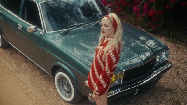 Blonde Woman In Retro Attire By Automobile. Serene Woman Resting Near Vehicle Amidst Lush Flowers