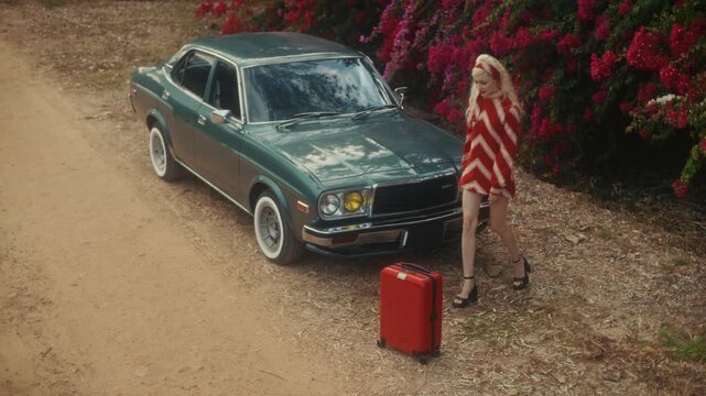 Blonde Woman In Retro Attire By Automobile. Serene Woman Resting Near Vehicle Amidst Lush Flowers
