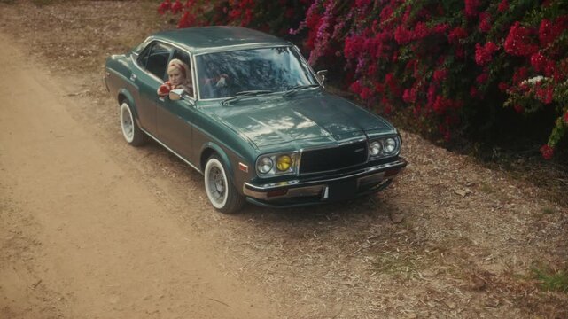 Blonde Woman In Retro Attire By Automobile. Serene Woman Resting Near Vehicle Amidst Lush Flowers