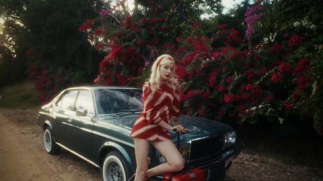 Blonde Woman In Retro Attire By Automobile. Serene Woman Resting Near Vehicle Amidst Lush Flowers