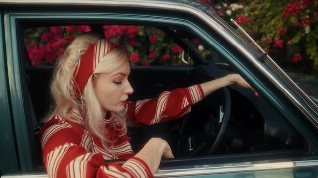 Blonde Woman In Retro Attire By Automobile. Serene Woman Resting Near Vehicle Amidst Lush Flowers