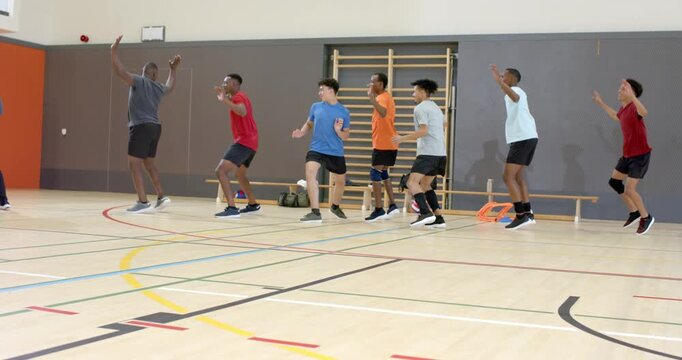 Diverse male athletes starting warmup at gym court lifting knees, jumping and rebounding with cones