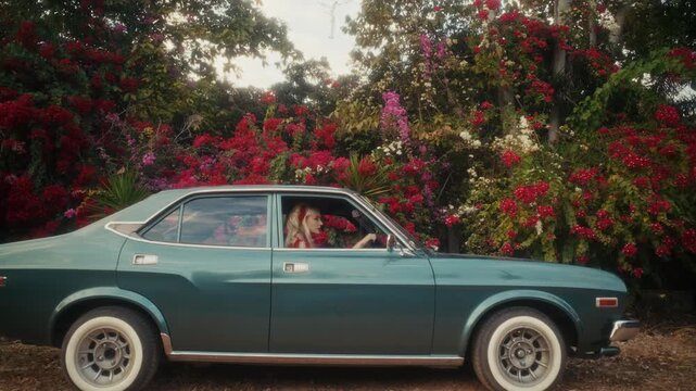 Blonde Woman In Retro Attire By Automobile. Serene Woman Resting Near Vehicle Amidst Lush Flowers