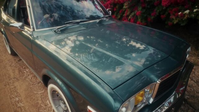 Blonde Woman In Retro Attire By Automobile. Serene Woman Resting Near Vehicle Amidst Lush Flowers