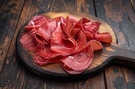 Thin cured beef slices bresaola meat arranged on a rustic wooden board with fresh rosemary over a dark textured background.