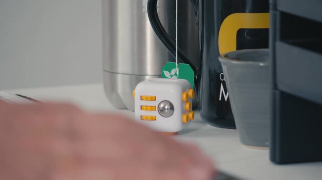 Small white fidget cube stress relief toy sitting on desk next to computer keyboard and mug. Modern workspace accessory for focus and anxiety management during work hours.