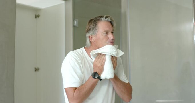 Mature man pressing and patting white towel on face while checking mirror after washing face