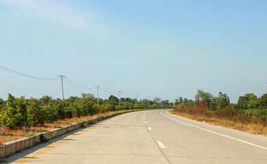 Long concrete road curves through sunny rural landscape bordered by green tropical bushes under a vast bright blue sky with utility poles.
