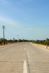 Empty concrete highway stretching into the distance under a bright blue sky, flanked by power poles and dry rural vegetation.