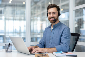 Fototapeta premium Smiling male call center agent with headset typing on laptop, providing friendly online customer support from a modern office desk, confident and professional portrait