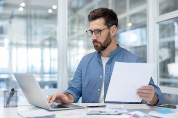 Fototapeta premium Confident businessman wearing glasses and a casual shirt working on his laptop, holding a report, and concentrating on analysis and data review in a modern office setup