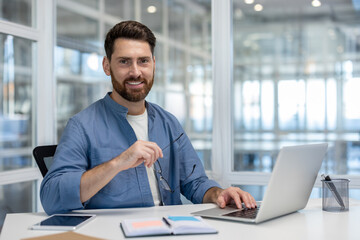 Fototapeta premium Confident bearded man smiling at the camera while working on a laptop at a bright desk, embodying professionalism, entrepreneurship, and productivity in a modern coworking space