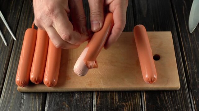 Hands peeling the skin off a hot dog sausage on a wooden cutting board with several sausages arranged in a row beside it