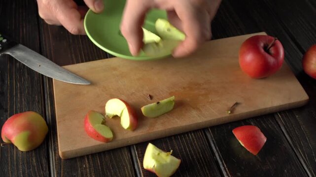 Hands placing sliced apples onto a green plate while preparing fruit on a wooden cutting board with a knife and whole apples in the background