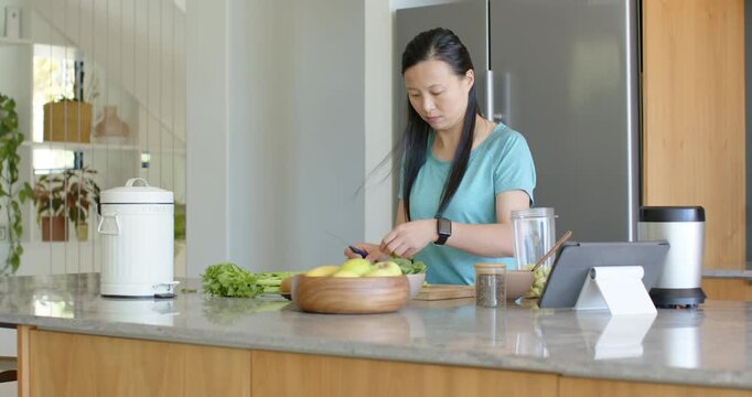 Asian woman in teal top chopping greens on island, tossing trimmings into white bin after piling