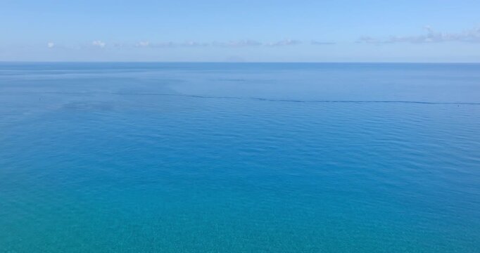Minimalist aerial view of the vast sea, showing a beautiful gradient from light turquoise near the shore to a deep sapphire blue, under a clear sky with a thin line of distant clouds.