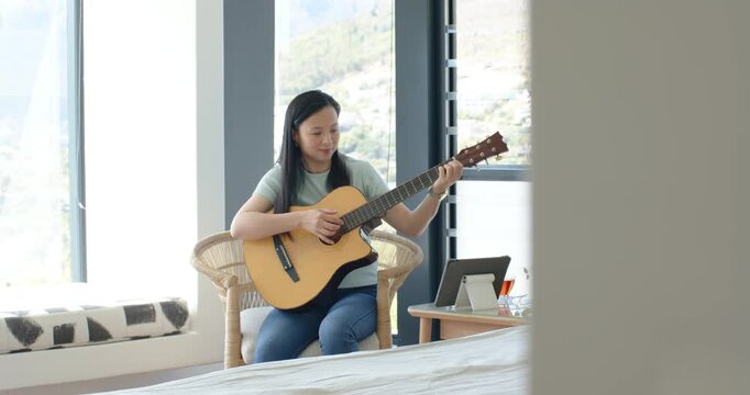 Asian woman sitting in wicker seat in bedroom by window practicing guitar following tablet lesson