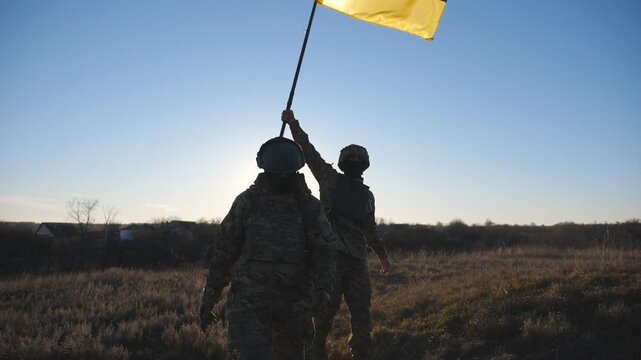 Female and male soldier of ukrainian army walking with raised flag of Ukraine on field at sunset. Woman and man in camouflage uniform going with blue-yellow banner at meadow. Invasion resistance