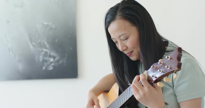 Asian woman positioning left-hand fingers, playing acoustic guitar at home, practicing chord phrase