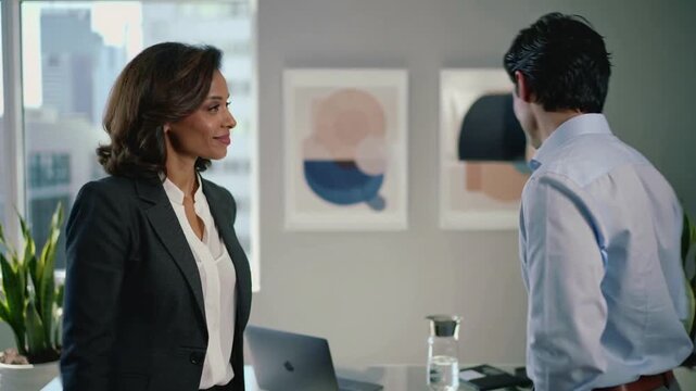 Stepping woman dark blazer offering handshake, man blue shirt shaking hands at desk ending meeting