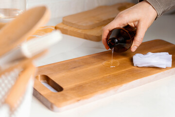 Hand pouring liquid oil from a dark glass bottle onto a wooden cutting board, with a cloth and...