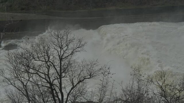 Massive Water Discharge from Dam with Nature Elements in Foreground. A brutal and powerful cascade of white rushing water discharging from a dam spillway. Close-up shot featuring tree branches 