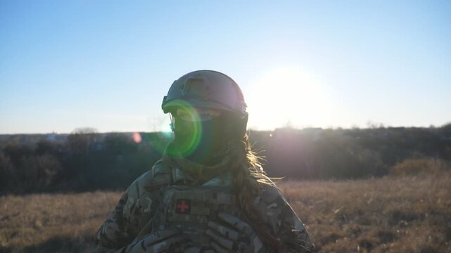 Young female military medic walking at field on sunny day. Ukrainian army woman in helmet and balaclava going on the meadow. Support against russian aggression. Concept of invasion resistance