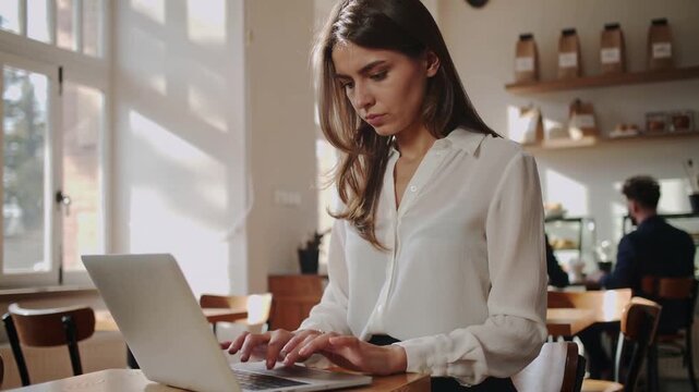 Working brown-haired woman wearing white blouse and dark pants typing on laptop at cafe, sunlight