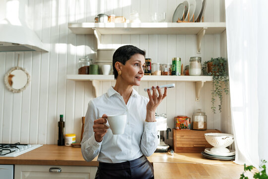 Successful classy female entrepreneur having coffee at her cozy kitchen while chatting with ai chat bot at voice mode, asking to process emails or summarize documents for current business projects
