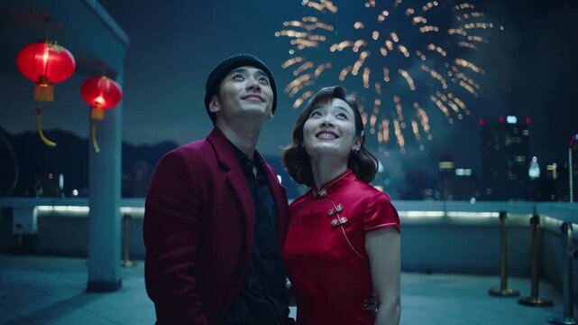 Smiling couple watching fireworks bursting over skyline on roof, red lanterns, beanie and cheongsam