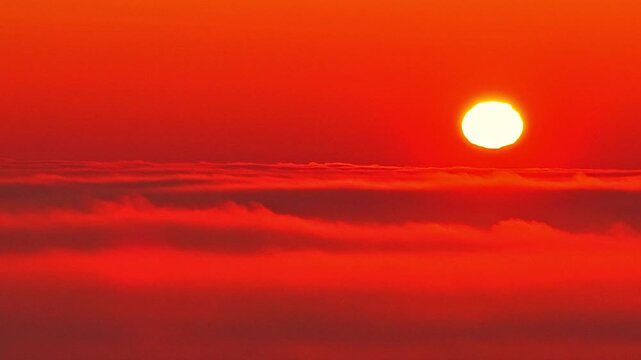 Drone view of the sun above a sea of clouds at dusk. Deep red tones fill the sky as sunlight fades along the horizon, creating a dramatic, abstract background with strong atmospheric mood.