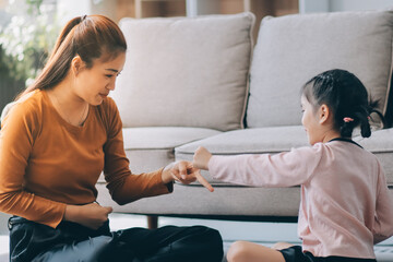 A cheerful Asian mother helps her young daughter do homework and color a book at home, while the father works on a laptop in the background. A cozy, bright, and modern family lifestyle.