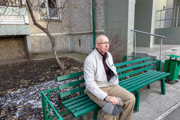 man with glasses sitting on a green bench outdoors in winter, concept of urban relaxation, waiting, or contemplation