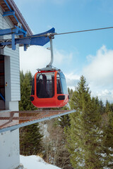 A red panoramic cable car gondola arrives at a mountain station platform surrounded by snow-covered pine trees under a blue sky