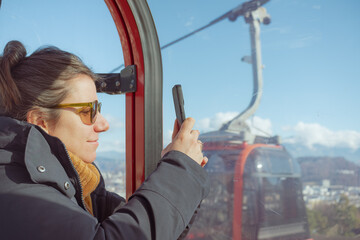 Young woman in a cable car gondola holding her smartphone to capture the panoramic mountain and city view during a sunny winter day © Pablo